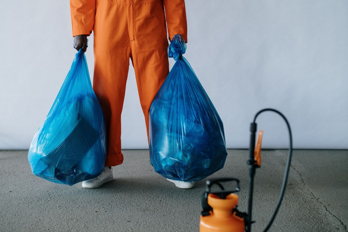 A cleaner in an orange uniform carrying two full trash bags