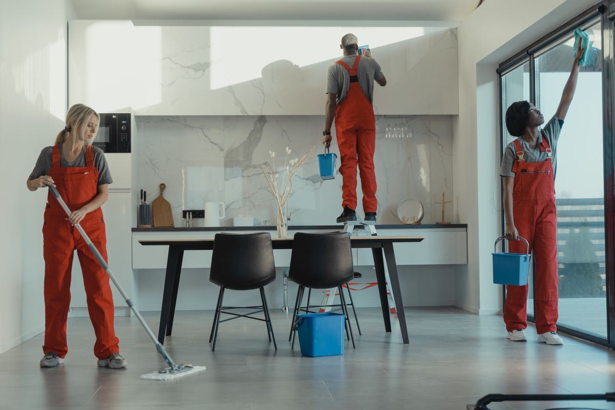 A three-person crew in red overalls mopping and cleaning windows in an office kitchen